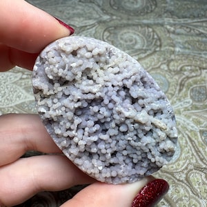 May include: An oval-shaped grape agate specimen featuring a cluster of small, spherical, light purple botryoidal crystals. The surface is densely covered in tiny, rounded, translucent purple formations that create a textured, bumpy appearance.