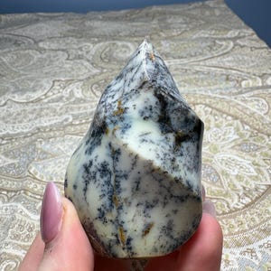 May include: A dendritic agate stone with a white base and black and brown tree-like inclusions. The polished stone has a smooth surface and a pointed top. The stone is held in a hand.