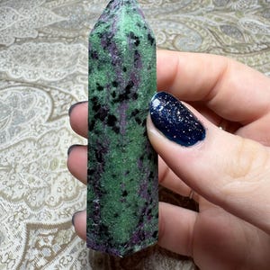 Może przedstawiać: Zielona i fioletowa wieża kryształowa zoisytu. Polerowany kamień ma prostokątny kształt ze spiczastym wierzchołkiem. Powierzchnia kryształu to mieszanka zieleni, fioletu i czerni, z nakrapianym wzorem. Kryształ trzymany jest w dłoni.