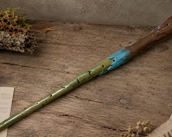 Op de afbeelding: Een handgemaakte toverstaf met een lange, taps toelopende olijfgroene schacht met kleine uitgesneden inkepingen, die overgaat in een heldere turquoise band en een gestructureerd, donkerbruin handvat met een knoestig, organisch uiterlijk.