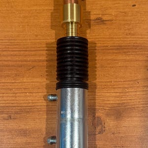 Puede incluir: Objeto cil&iacute;ndrico met&aacute;lico con una parte superior roscada y una serie de componentes. Presenta una base plateada, una secci&oacute;n negra acanalada y detalles en cobre y lat&oacute;n. El objeto est&aacute; sobre una superficie de madera.