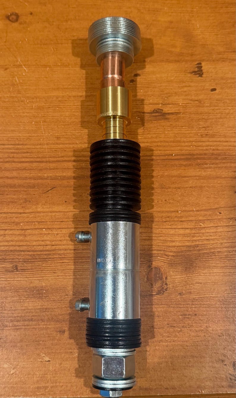 Puede incluir: Objeto cil&iacute;ndrico met&aacute;lico con una parte superior roscada y una serie de componentes. Presenta una base plateada, una secci&oacute;n negra acanalada y detalles en cobre y lat&oacute;n. El objeto est&aacute; sobre una superficie de madera.