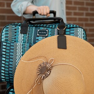 May include: A blue and white patterned rolling suitcase with a brown straw sun hat on top. The suitcase has a black handle and a black luggage tag with the text "Vera Bradley" on it.