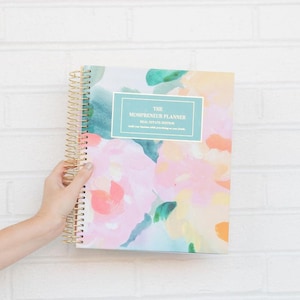 May include: A spiral-bound planner with a floral design in pink, yellow, and green hues. The cover features the text "THE MOMPRENEUR PLANNER REAL ESTATE EDITION". The planner has a gold spiral binding.