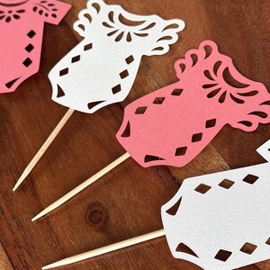 May include: A collection of die-cut paper baby onesie toppers in pink and white, each featuring intricate floral and diamond-shaped cutouts. Every paper cutout is securely attached to a thin, pointed wooden toothpick for easy insertion into food.