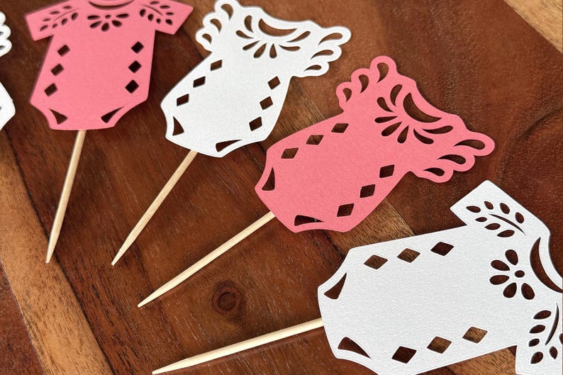 May include: A collection of die-cut paper baby onesie toppers in pink and white, each featuring intricate floral and diamond-shaped cutouts. Every paper cutout is securely attached to a thin, pointed wooden toothpick for easy insertion into food.