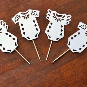 May include: Four white paper cutouts shaped like baby bodysuits, each attached to a thin wooden toothpick. The paper cutouts feature intricate die-cut patterns including floral motifs around the neckline and a series of diamond shapes on the torso.