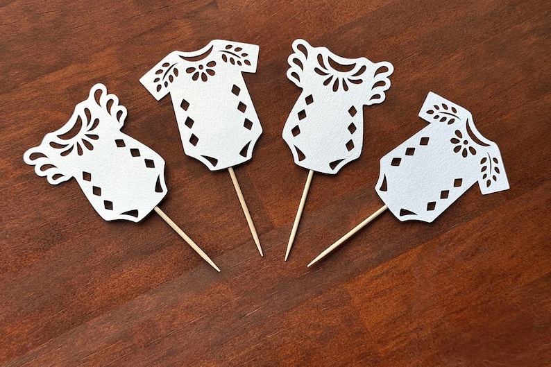 May include: Four white paper cutouts shaped like baby bodysuits, each attached to a thin wooden toothpick. The paper cutouts feature intricate die-cut patterns including floral motifs around the neckline and a series of diamond shapes on the torso.