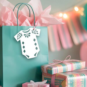 May include: A teal gift bag filled with light pink tissue paper, featuring a white die-cut tag shaped like a baby onesie with floral and diamond patterns. The bag is surrounded by several wrapped gifts in teal and pink patterned paper.