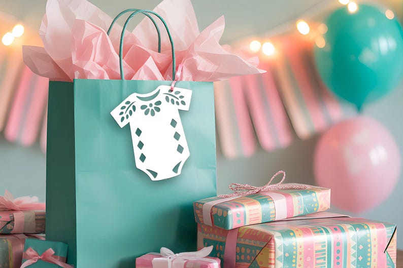 May include: A teal gift bag filled with light pink tissue paper, featuring a white die-cut tag shaped like a baby onesie with floral and diamond patterns. The bag is surrounded by several wrapped gifts in teal and pink patterned paper.