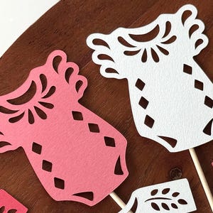 May include: A collection of die-cut paper baby onesie shapes in pink and white, featuring intricate cutout patterns around the neckline and diamond-shaped cutouts on the body, each attached to a thin wooden stick for use as a decorative topper.