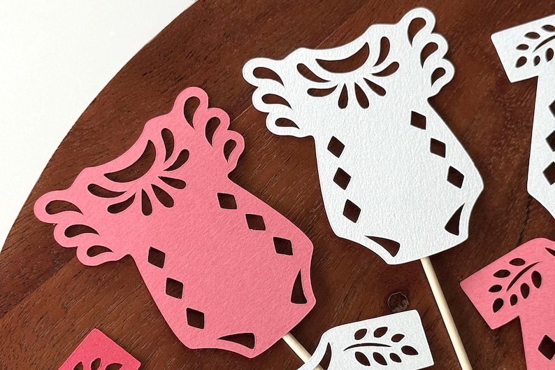 May include: A collection of die-cut paper baby onesie shapes in pink and white, featuring intricate cutout patterns around the neckline and diamond-shaped cutouts on the body, each attached to a thin wooden stick for use as a decorative topper.