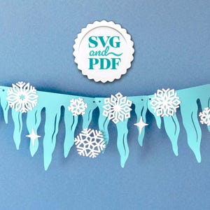 Peut inclure: Une guirlande en papier bleu clair avec des flocons de neige et des glaçons blancs découpés, tendue sur une fine ficelle blanche. La bannière comporte également des découpes d'étoiles blanches. Un logo circulaire blanc avec le texte "SVG and PDF" est en haut.