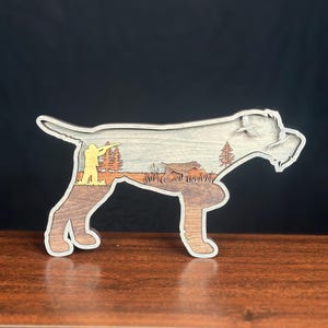 Puede incluir: Un recorte de madera multicapa con forma de perro de caza. El interior presenta una escena cortada con láser con un cazador amarillo apuntando un rifle, un perro marrón en hierba alta, pinos y un pájaro volando sobre un fondo gris claro.
