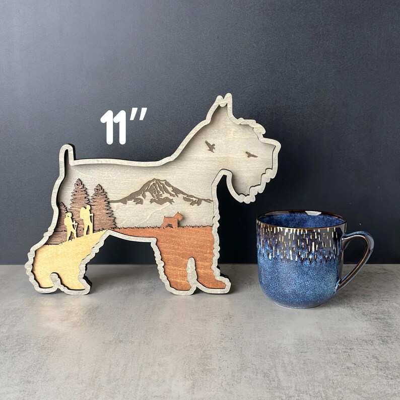 Puede incluir: Pieza de arte de madera con forma de perro con una escena de monta&ntilde;a y bosque en el interior. La pieza de arte mide 28 cm de largo. Una taza de cer&aacute;mica azul y marr&oacute;n est&aacute; al lado de la pieza de arte de madera. El fondo es de color gris oscuro.