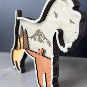 Puede incluir: Silueta de madera en capas de un perro Schnauzer con una escena de monta&ntilde;a y bosque. El perro es marr&oacute;n, beige y en tonos de madera natural, con un contorno marr&oacute;n oscuro. Dos p&aacute;jaros vuelan en el cielo. Es un objeto decorativo.