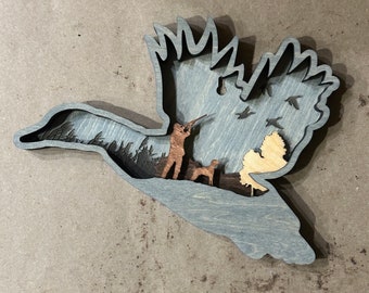 May include: A layered wooden art piece in the shape of a flying duck. The duck is gray with a brown hunter and dog inside. Additional wooden elements include trees, birds, and a light-colored wing.