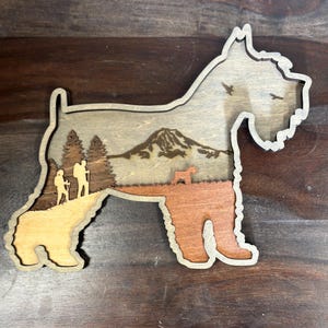 Puede incluir: Recorte de madera con forma de perro y dise&ntilde;o en capas. El interior presenta una escena de monta&ntilde;a con excursionistas y &aacute;rboles. Las patas del perro son de color marr&oacute;n anaranjado c&aacute;lido, mientras que el cuerpo es de tono madera natural. Dos p&aacute;jaros vuelan en el cielo.