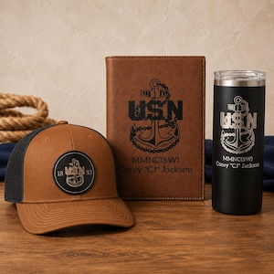 Op de afbeelding: Een bruine en zwarte honkbalpet met een ronde patch met een anker en de tekst USN 18 93, een bruin notitieboek in lederlook met een anker en USN MMNC(SW) Corey CJ Jackson, en een zwarte beker met hetzelfde anker en dezelfde tekst.