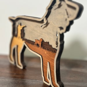 以下が含まれることがあります： 木製の犬のシルエット彫刻。明るい茶色、中茶色、濃い茶色の配色です。犬の形の内側には、木や他の犬がいる風景が描かれています。彫刻は濃い茶色の表面に立っています。