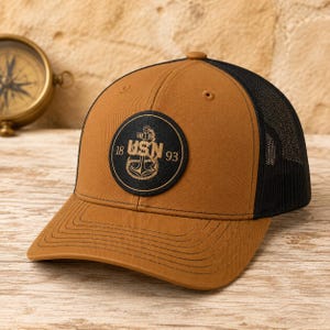 May include: A tan baseball cap with a black mesh back and a circular black patch on the front. The patch features a gold anchor graphic, the text USN, and the numbers 18 and 93. The cap has a curved brim with visible stitching detail.