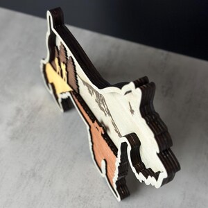 Puede incluir: Una obra de arte de madera multicapa que representa un paisaje monta&ntilde;oso. Presenta tonos de marr&oacute;n, beige y blanco, con un contorno marr&oacute;n oscuro. El dise&ntilde;o muestra una detallada cordillera y una silueta estilizada de animal.