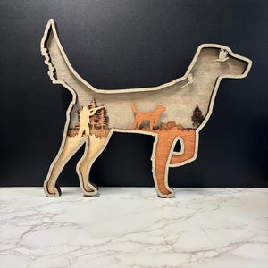 Puede incluir: Una pieza de arte mural de madera multicapa con forma de perro de caza. El recorte interior presenta una escena con un cazador apuntando con un rifle, pinos, la silueta de un perro más pequeño y un ave en vuelo, todo en tonos de madera natural.