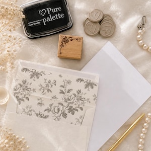 May include: An assortment of stationery supplies, including a black ink pad with "Pure palette" text, a wooden stamp, heart-shaped wax seals, envelopes, a gold pen, and a pearl necklace. The envelopes have a floral design.