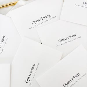 Puede incluir: Un conjunto de tarjetas blancas con texto negro, dispersas sobre una superficie blanca. Cada tarjeta presenta las palabras "Open when" o "Open during" seguidas de una frase corta. Una idea de regalo reflexiva.