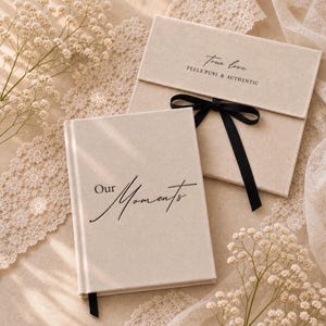 May include: A beige hardcover book with the text Our Moments in black script, accompanied by a matching beige presentation box featuring a black ribbon tie and the text True love FEELS PURE & AUTHENTIC printed in black on the front flap.