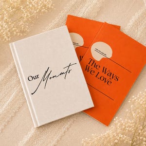May include: Three notebooks, two orange and one off-white, are arranged on a textured surface. The off-white notebook has the words "Our Moments" in black script. The orange notebooks read "The Ways We Love" and "For You and Me."