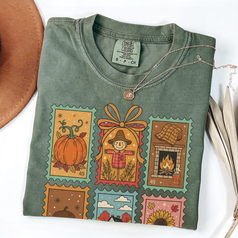 Puede incluir: Camiseta verde Comfort Colors con un dise&ntilde;o de sellos coloridos con una calabaza, un espantap&aacute;jaros, una chimenea y un girasol. Un collar plateado con un colgante descansa sobre la camisa. Un sombrero marr&oacute;n es visible en el fondo.