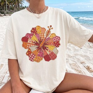 May include: Cream-colored t-shirt featuring a large, colorful hibiscus flower design. The flower is composed of various fabric patches in shades of red, yellow, orange, and pink. The shirt has a relaxed fit and short sleeves, perfect for casual wear.