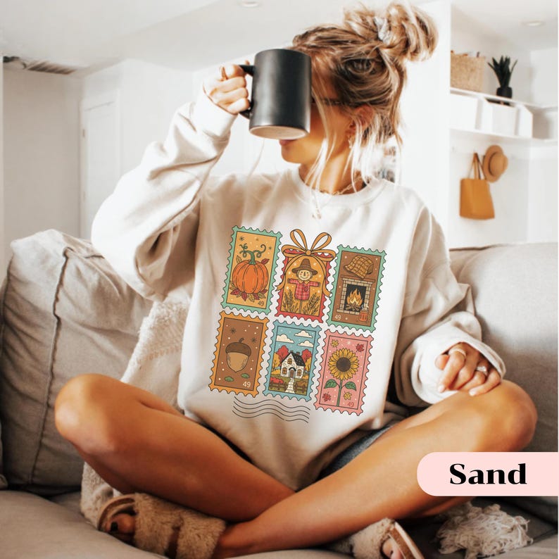 Puede incluir: Una sudadera de color arena con un dise&ntilde;o de sellos coloridos con ilustraciones de tem&aacute;tica oto&ntilde;al. Los sellos representan una calabaza, un espantap&aacute;jaros, una chimenea, una bellota, una casa y un girasol. La palabra "Sand" est&aacute; impresa en la parte inferior.