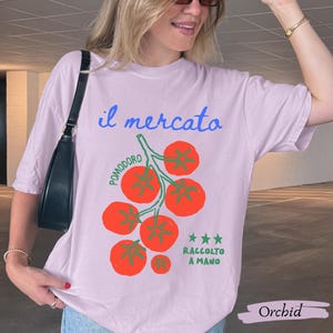 May include: Orchid-colored t-shirt with the text "il mercato" in blue script above a graphic of red tomatoes on a green vine. The text "POMODORO" is arched above the tomatoes, and "RACCOLTO A MANO" is below. The shirt is a relaxed fit.