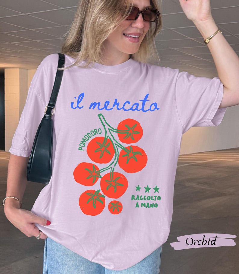 May include: Orchid-colored t-shirt with the text "il mercato" in blue script above a graphic of red tomatoes on a green vine. The text "POMODORO" is arched above the tomatoes, and "RACCOLTO A MANO" is below. The shirt is a relaxed fit.