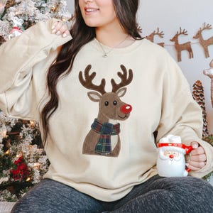 May include: A cream-colored crewneck sweatshirt featuring a central graphic of a brown reindeer with a textured appearance, large antlers, a bright red nose, and a green, blue, and red plaid scarf wrapped around its neck.