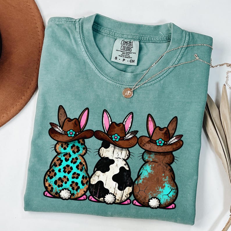 May include: A teal Comfort Colors t-shirt featuring three cartoon bunnies wearing cowboy hats. The bunnies have different patterns: leopard print, black and white cow spots, and brown with turquoise accents. A silver necklace with a pendant is also visible.
