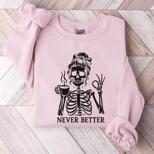 May include: A pale pink sweatshirt featuring a black skeleton graphic with a bun, holding a coffee cup and giving an okay sign. The text "NEVER BETTER" is below the skeleton. The sweatshirt is folded on a light wood grain surface.