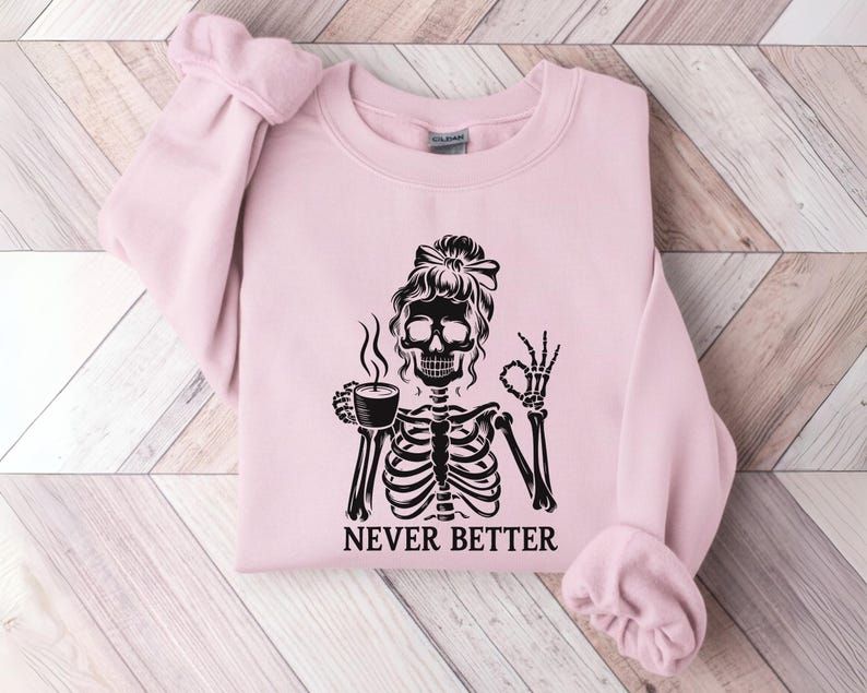 May include: A pale pink sweatshirt featuring a black skeleton graphic with a bun, holding a coffee cup and giving an okay sign. The text "NEVER BETTER" is below the skeleton. The sweatshirt is folded on a light wood grain surface.