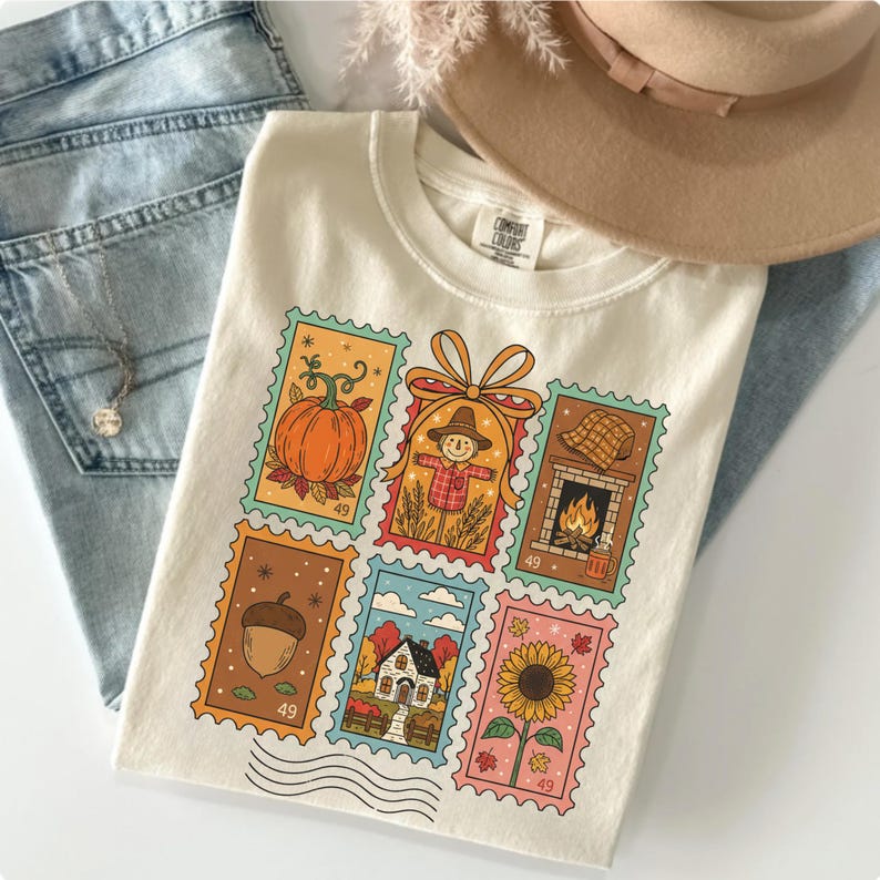 Puede incluir: Camiseta color crema con dise&ntilde;os de sellos postales coloridos con temas oto&ntilde;ales. Los sellos representan una calabaza, un espantap&aacute;jaros, una chimenea, una bellota, una casa y un girasol. La camiseta incluye el texto "COMFORT COLORS".