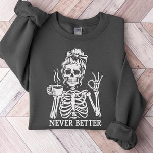 May include: Dark grey sweatshirt with a white skeleton graphic. The skeleton, with a bow in its hair, holds a coffee cup and makes an "okay" hand gesture. The text "NEVER BETTER" is printed below the graphic.