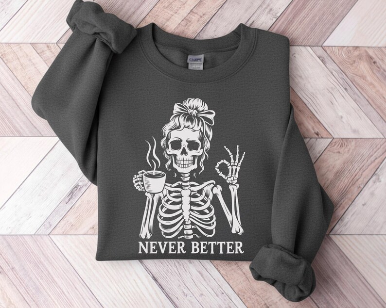 May include: Dark grey sweatshirt with a white skeleton graphic. The skeleton, with a bow in its hair, holds a coffee cup and makes an "okay" hand gesture. The text "NEVER BETTER" is printed below the graphic.