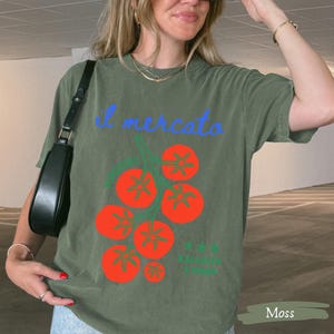 May include: Olive green t-shirt with the Italian text "il mercato" in blue script above a graphic of red tomatoes. The shirt has a relaxed fit and short sleeves. The word "Moss" is in a rectangle at the bottom.