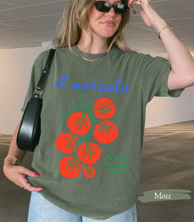May include: Olive green t-shirt with the Italian text "il mercato" in blue script above a graphic of red tomatoes. The shirt has a relaxed fit and short sleeves. The word "Moss" is in a rectangle at the bottom.