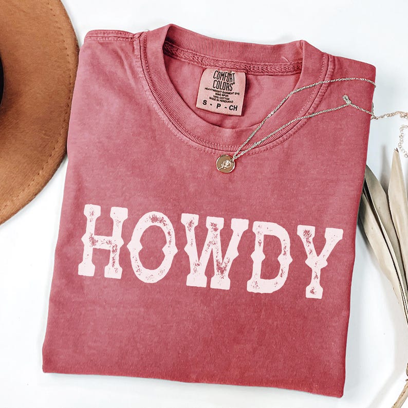 May include: A faded rose-coloured t-shirt with the word "HOWDY" in distressed white lettering. The shirt has a crew neck and a small tag at the neckline. A silver necklace with a round pendant is draped across the shirt. A brown hat is partially visible.