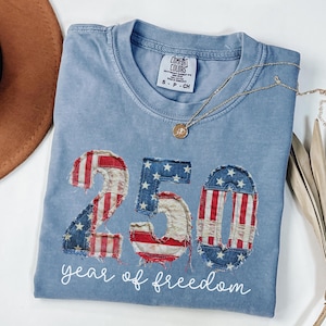 Puede incluir: Una camiseta azul claro con el número "250" en un diseño de bandera estadounidense desgastada. El texto "year of freedom" está debajo. Un collar dorado con un colgante está sobre la camiseta.