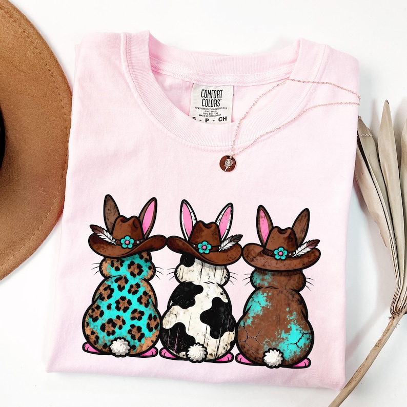 May include: A light pink t-shirt featuring three cartoon bunnies wearing cowboy hats. The bunnies have different patterns: leopard print, black and white cow print, and brown with turquoise accents. A gold necklace and a brown hat are also visible.