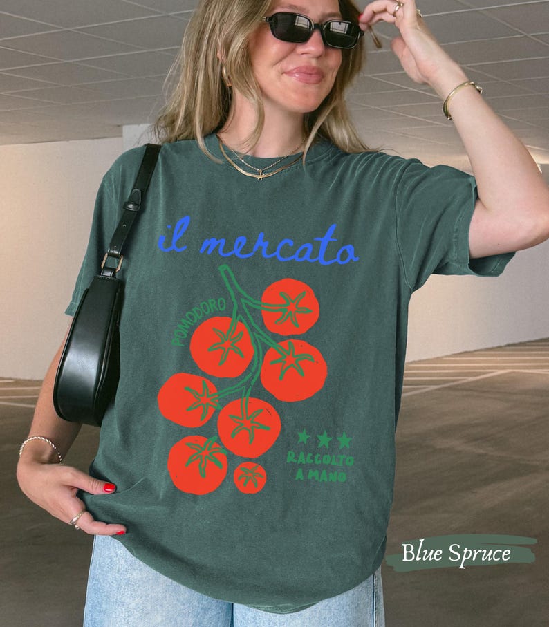 May include: Blue spruce t-shirt with a tomato graphic. The design includes the words "il mercato" in blue script, a cluster of red tomatoes, and the words "RACCOLTO A MANO".