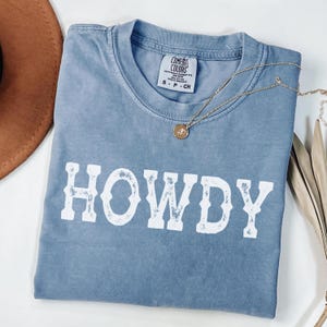 May include: A faded blue Comfort Colors t-shirt with the word "HOWDY" in white block letters. A gold necklace with a pendant rests on the shirt. A brown hat is visible in the upper left corner.